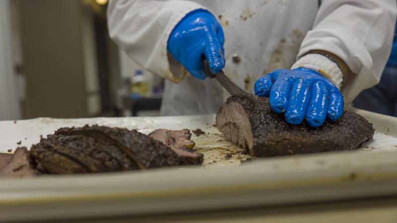 A person wearing blue gloves slices up meat. 